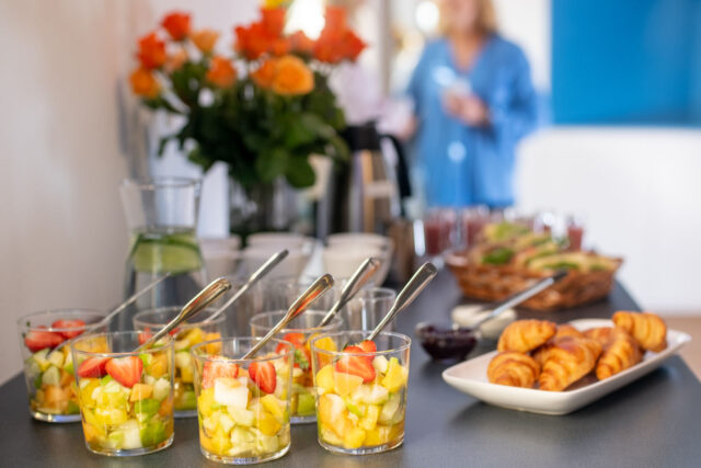 Fruit salad in dessert bowls is the centre of attention, with croissants neatly laid out on a plate just behind and orange flowers in a blurred background.