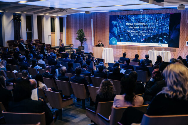 An audience listens to the speaker on stage. The speaker has a large screen behind them with a bulleted list for the audience to follow.