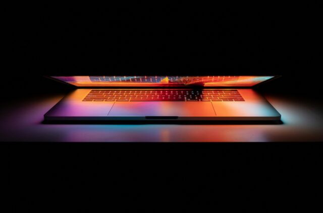 en bärbar dator med skärmen halvt nere och färgglatt ljus som reflekteras över tangentbordet mot den mörka bakgrunden.