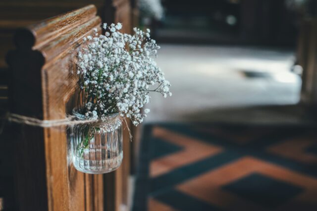 Bröllopsdekoration i kyrkan i form av en glasburk med växten brudslöja i, knuten runt yttre delen av en kyrkbänk.