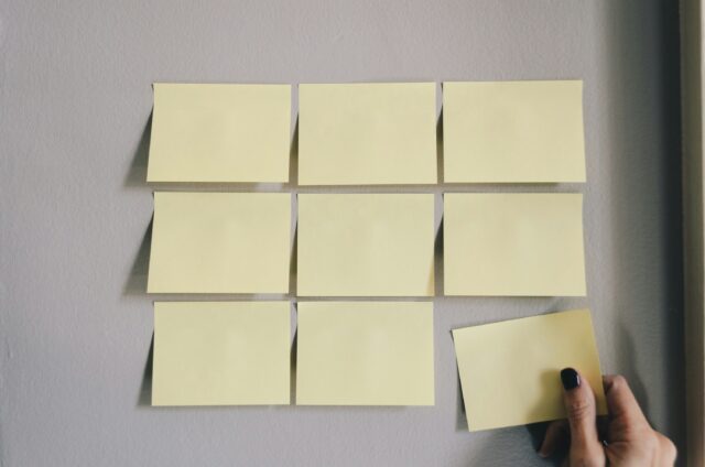Nio gula post-it-lappar hänger i ett rutnät på en vägg med en hand som tar ner den nionde.